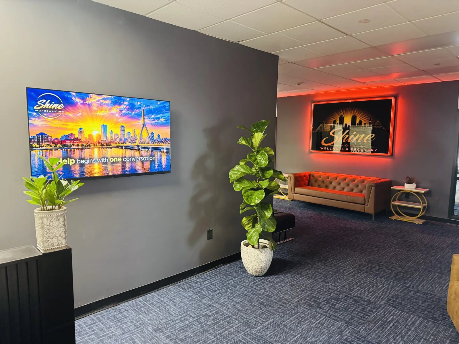 Reception lounge at Shine Wellness & Recovery with a wall-mounted TV showing the Shine Boston sunset welcome screen, potted plants, and the illuminated Shine sign glowing red in the background