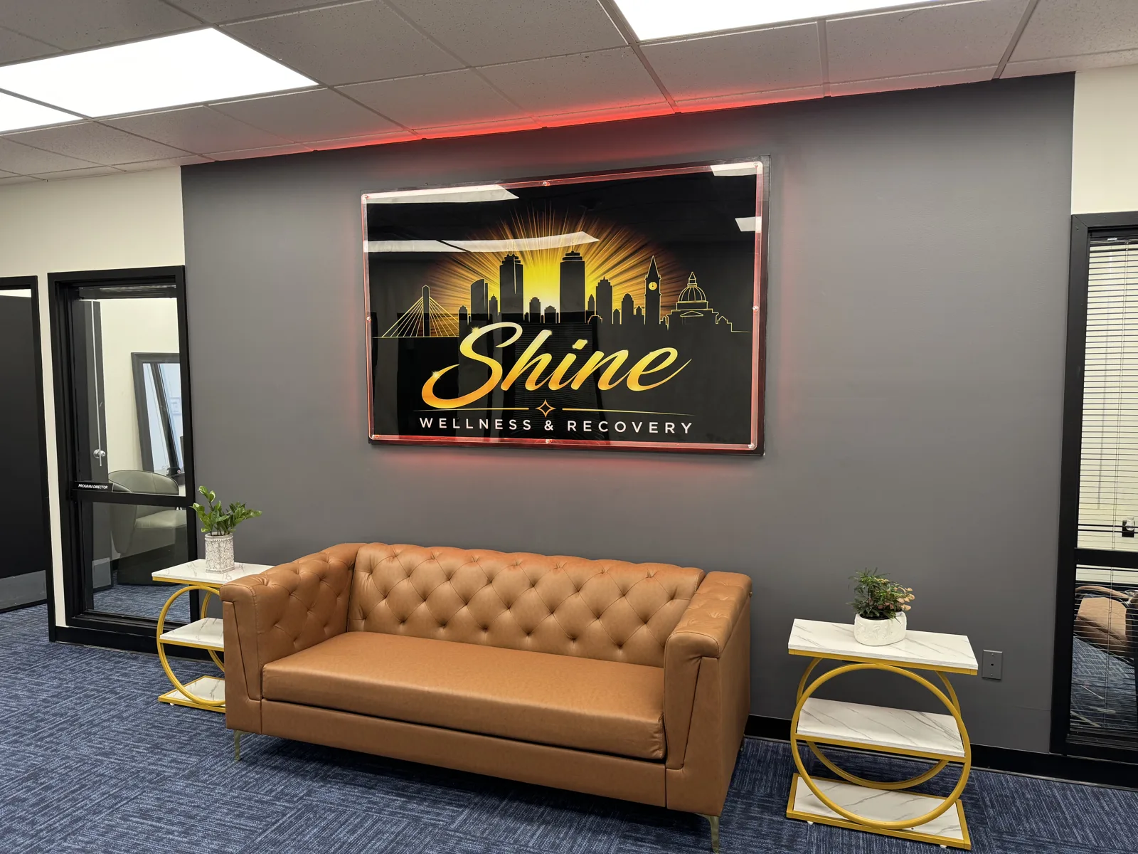Lounge at Shine Wellness & Recovery with a tan leather Chesterfield sofa, gold marble side tables, and an illuminated Shine Wellness & Recovery sign on a dark gray wall