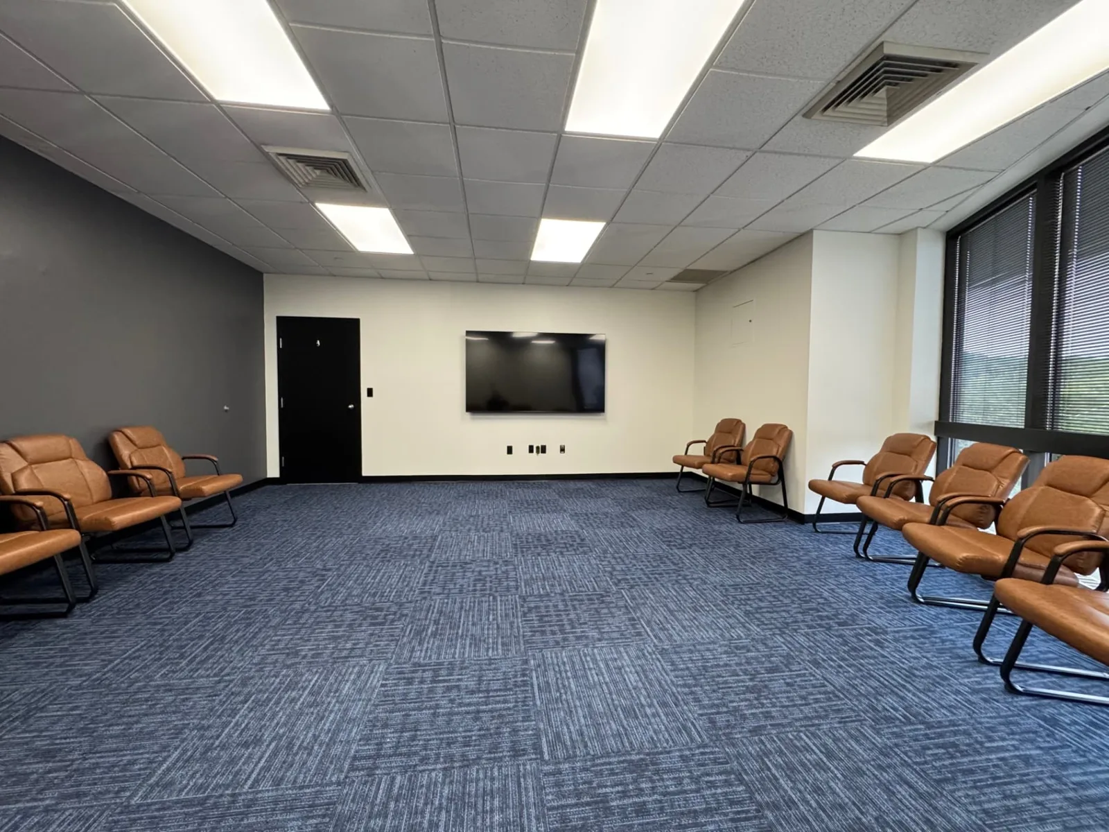 Main group therapy room at Shine Wellness & Recovery with tan chairs arranged in a circle, wall-mounted TV, and window wall