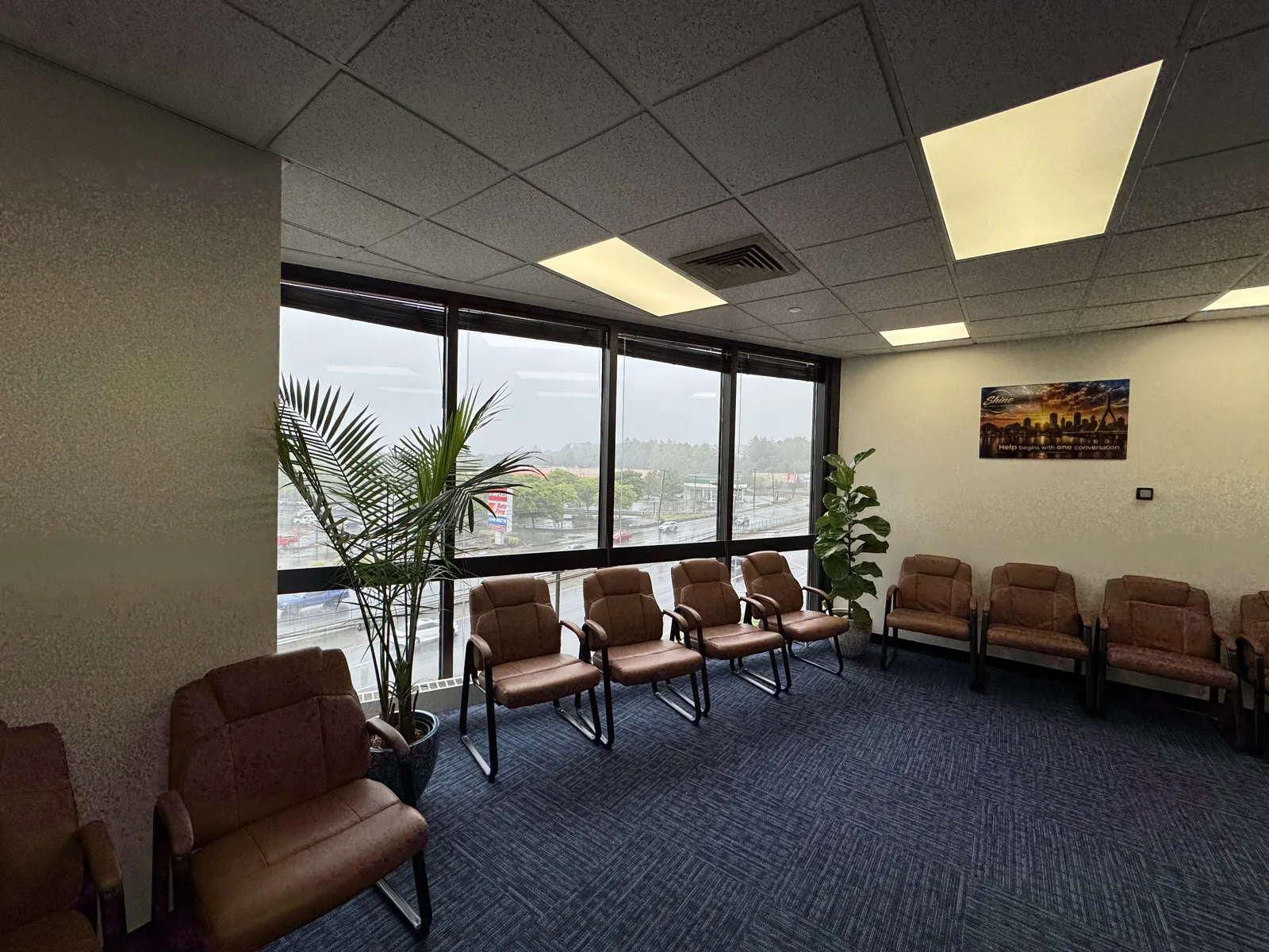 Group room at Shine Wellness & Recovery with two rows of brown leather chairs along windows overlooking Broadway, potted plants, and a Shine canvas print that reads Help begins with one conversation